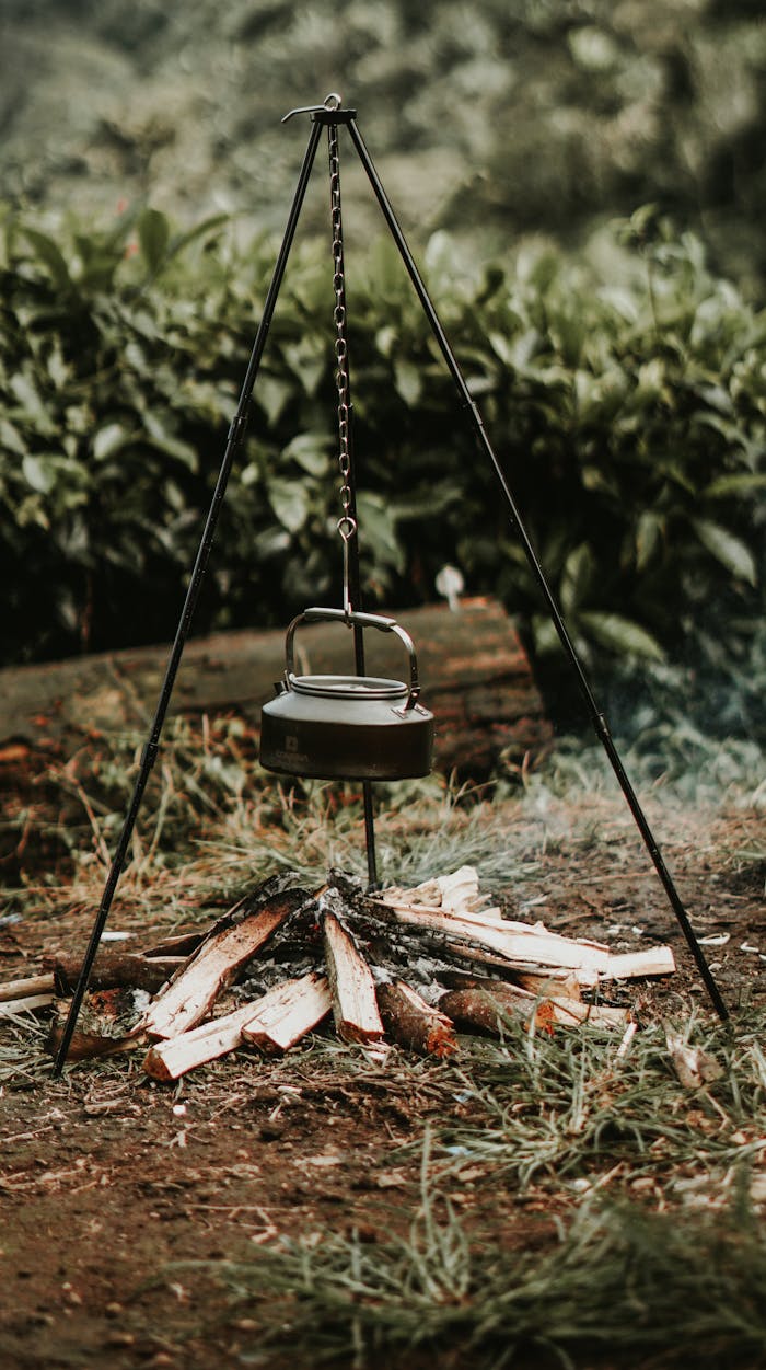 Kettle over a campfire tripod amidst West Java's lush forest, capturing a rustic camping vibe.